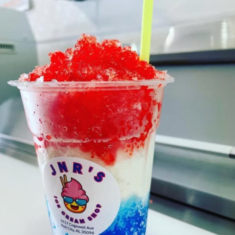 A JNR's Ice Cream Shop cup filled with red, white, and blue shaved ice and a yellow straw.