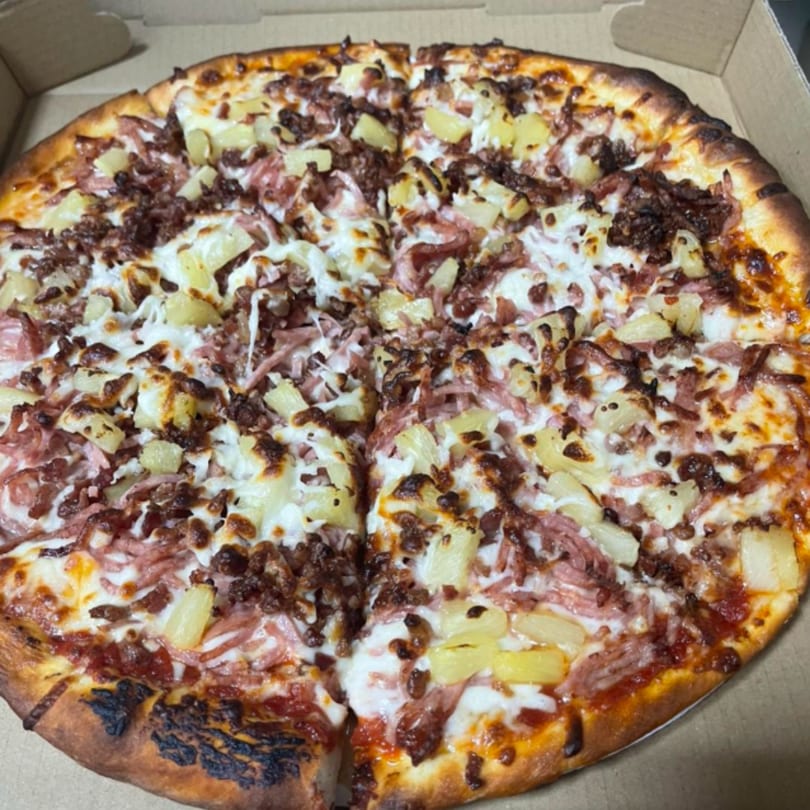 A whole JNR's pizza with pineapple, ham, and bacon, cut into slices in a cardboard box.