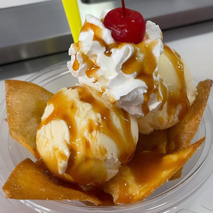 Two scoops of vanilla ice cream with caramel, whipped cream, and a cherry in a clear bowl.