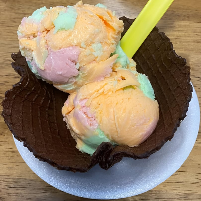 Two scoops of colorful ice cream in a chocolate waffle bowl with a yellow spoon.